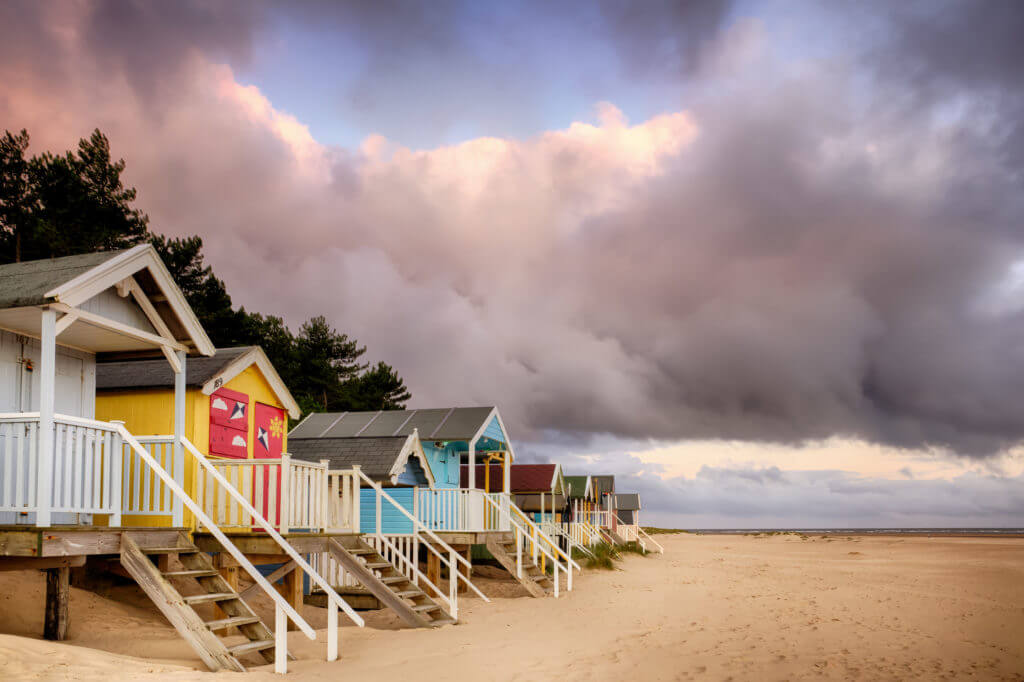Elevated beach huts in Norfolk are some of the most colourful in England, situated on the sandy North Norfolk coast. Sunrise clouds with an early morning pink tint. Steps leading down the the sandy shoreline from the wooden beach huts. Norfolk has been awarded the most 'Blue Flags' for high quality beaches in the country.
