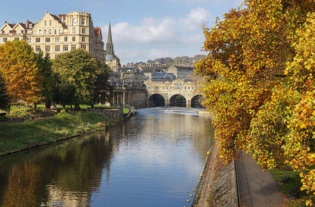 The famous landmarks of Pulteney Weir and Pulteney Bridge surrounded by vibrant autumnal trees on the River Avon in Bath, UK.