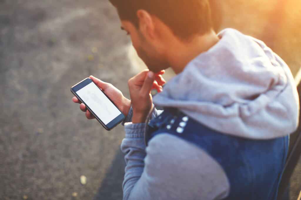 Close up handsome young man using smart phone while standing outdoors at sunny evening, rear view shot hipster man holding cell phone while looking to some fashion website on mobile screen, flare