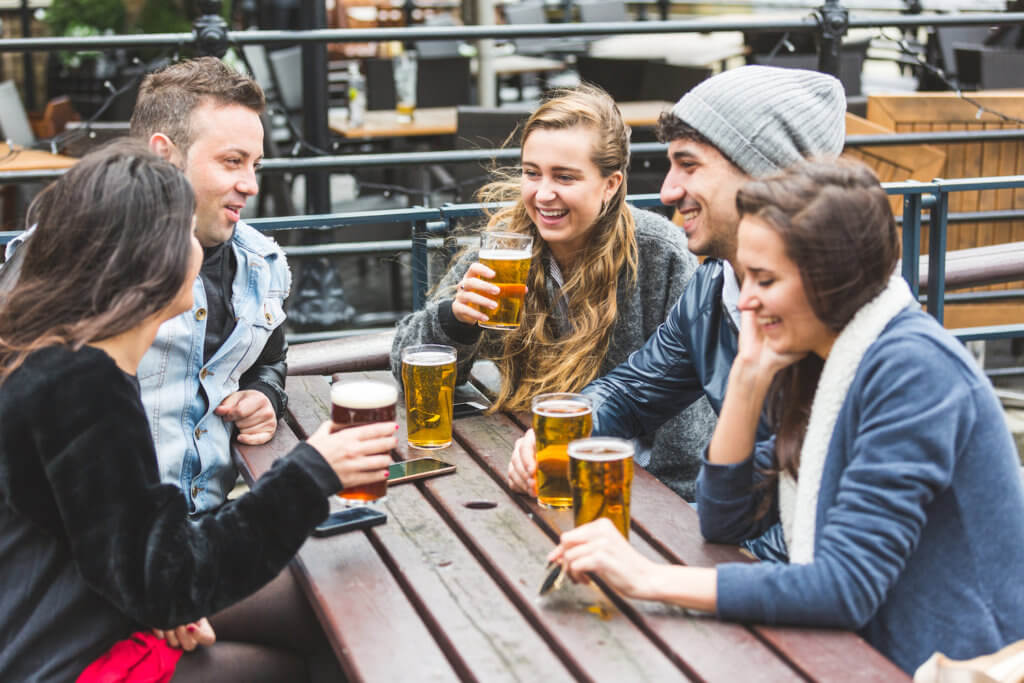 Group of friends enjoying a beer at pub in London, toasting and laughing. They are seated outside at a wood table, wearing winter clothes. Friendship and lifestyle concepts.